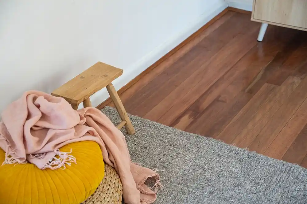 Ahşap zeminli odada sarı koltuk üzerinde pembe battaniye, yanında ahşap tabure ve gri halı
